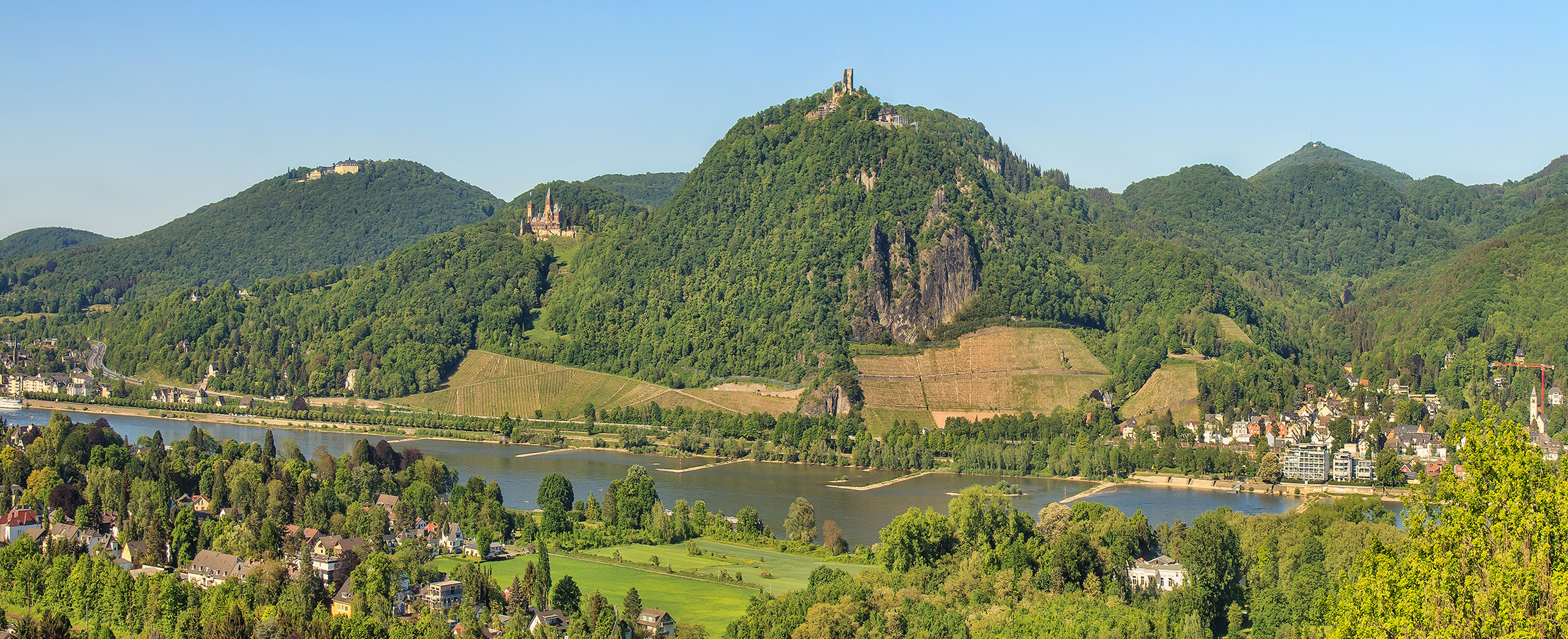 Blick vom Rheintal auf das Siebengebirge mit der Burgruine Drachenfels und Schloss Drachenburg bei strahlendem Sonnenschein.