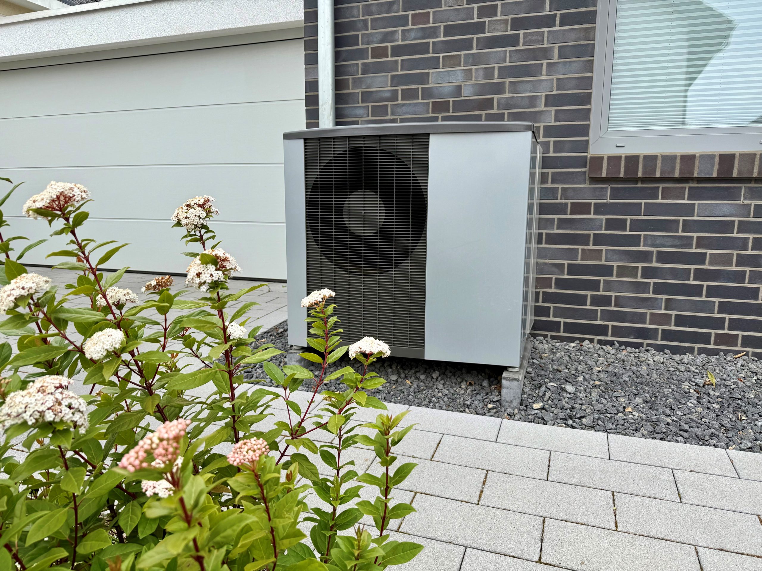 Moderne Luft-Wasser-Wärmepumpe vor modernem Haus mit Blumen im Vordergrund.