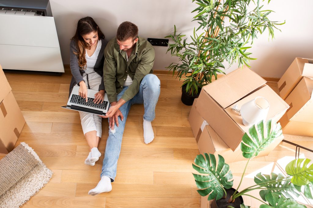 Mann und Frau sitzen auf dem Boden mit dem Laptop in der Hand. Neben beiden stehen gepackte Umzugskartons.