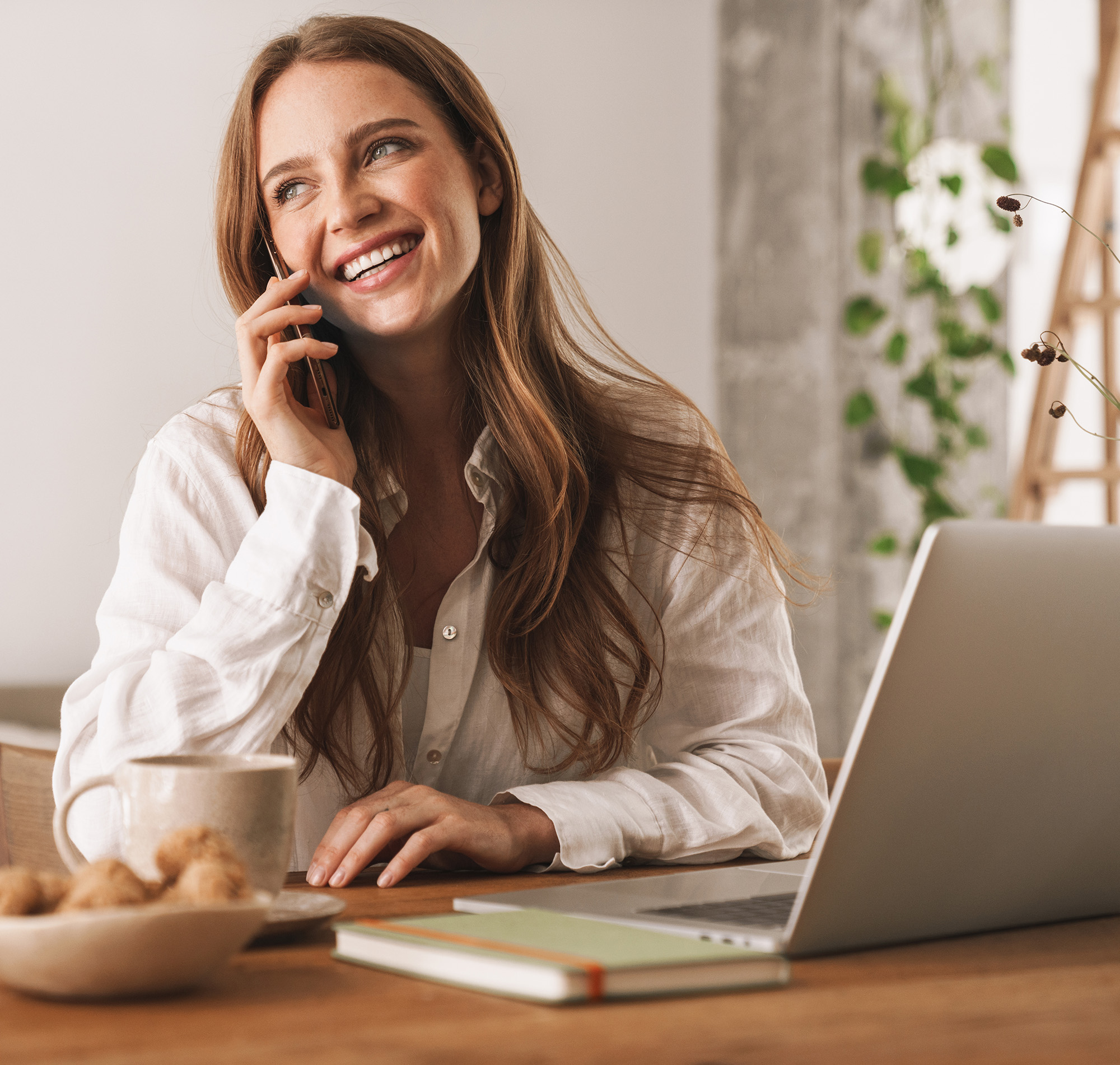 Frau sitzt vor dem Laptop und telefoniert mit dem Smartphone.